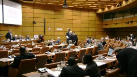 The picture shows the meeting room, which has light brown wooden paneled walls and brown leather chairs. In the picture are meeting attendees in discussion.